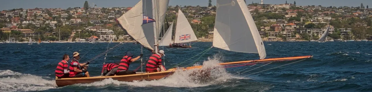 Crew in red-and-black hooped jerseys driving an 18-footer through a wave, Britannia's Union Jack sail astern