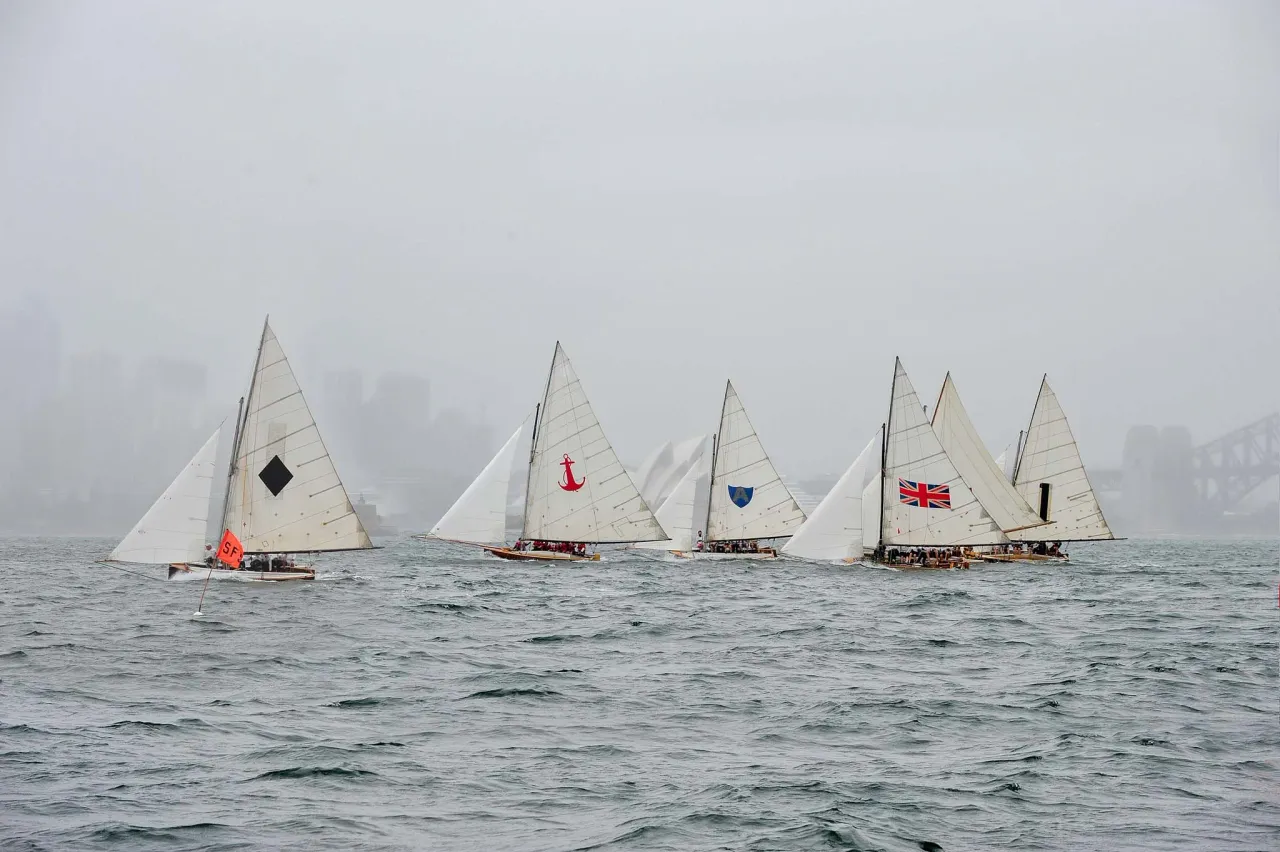 Historic 18-footers carrying diamond, anchor, shield and Union Jack insignia ghosting past the Opera House in rain