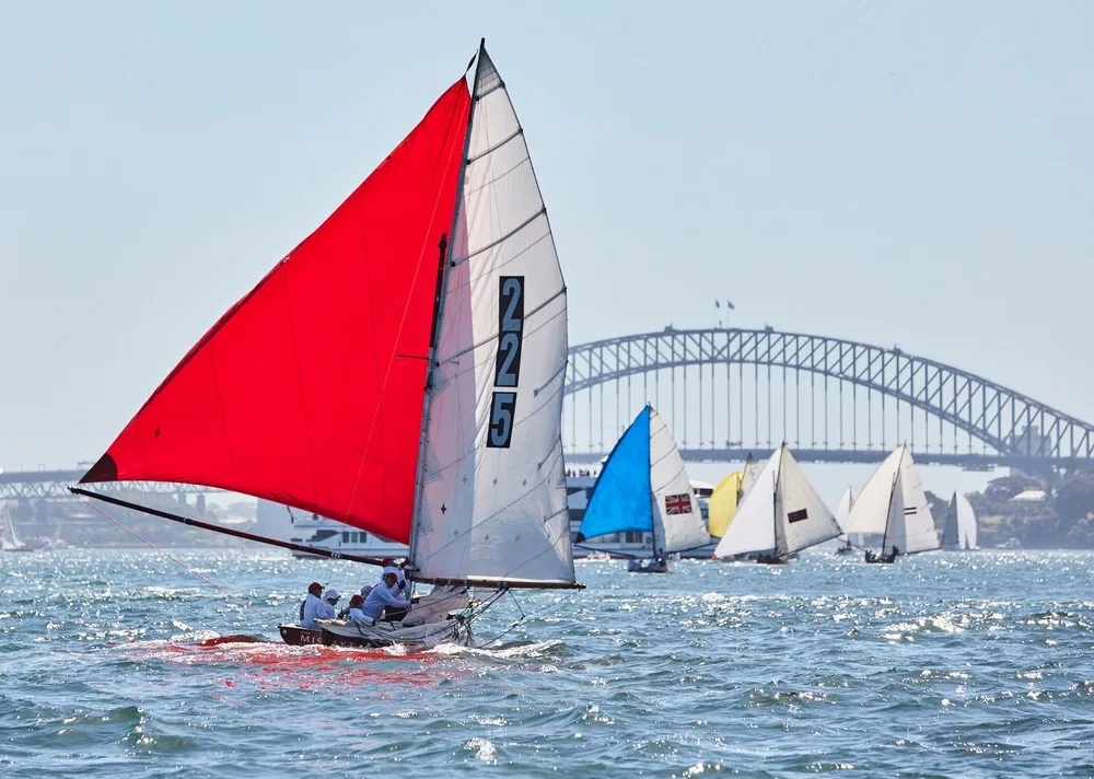Historic 18-footer skiffs with red, blue and yellow sails racing in front of the Sydney Harbour Bridge