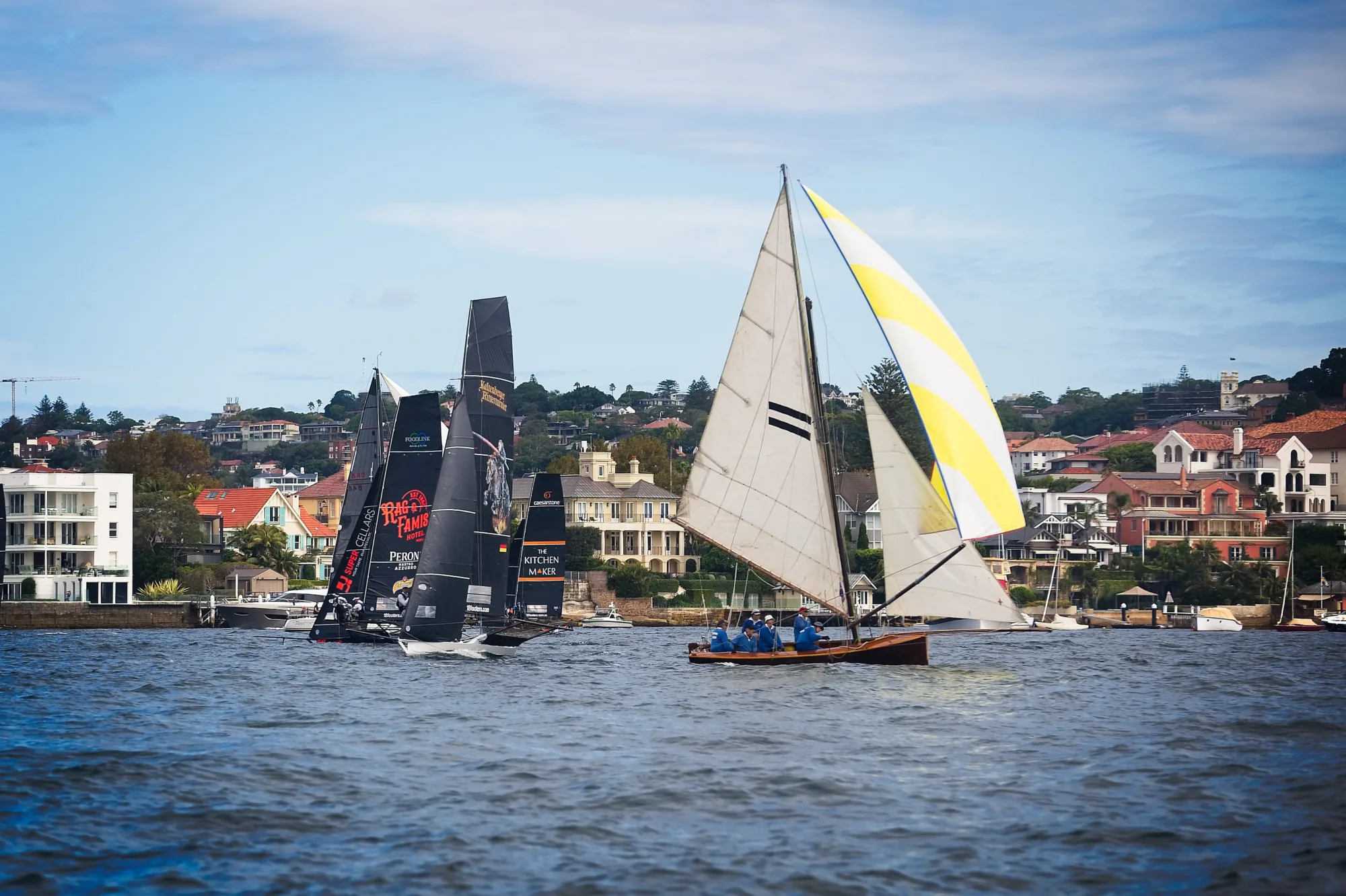 The replica Tangalooma racing under a yellow and white spinnaker, two horizontal black bars on the mainsail, with modern 18-footers astern